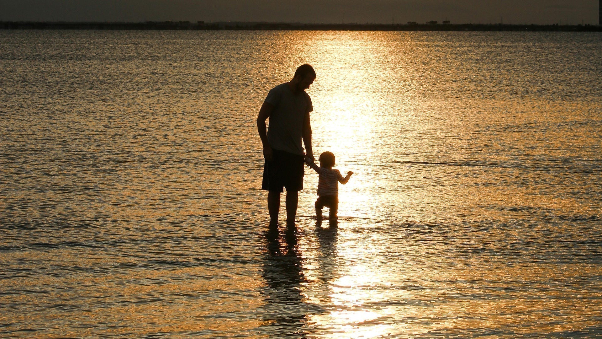 Silhouette of a father holding his child’s hand while walking through shallow water at sunset – warm golden reflections over a peaceful seascape.