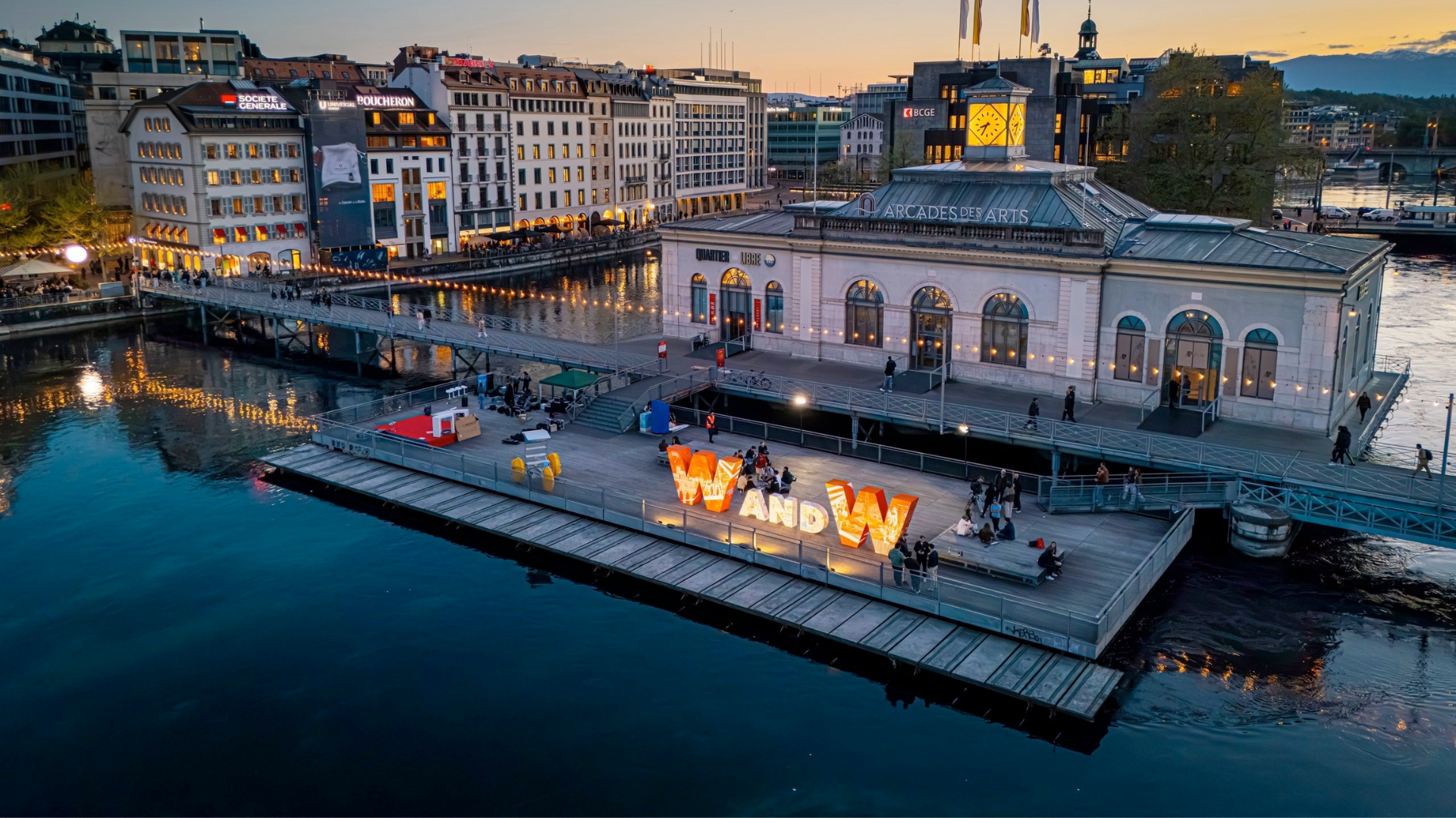 Evening view of Geneva’s waterfront with illuminated buildings and the Watches & Wonders installation along the lake.